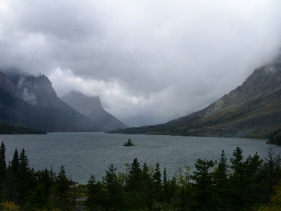 [A tiny island in the middle of a large round lake surrounded by mountains on the sides and back with lots of clouds overhead and blocking the view of some mountains.]